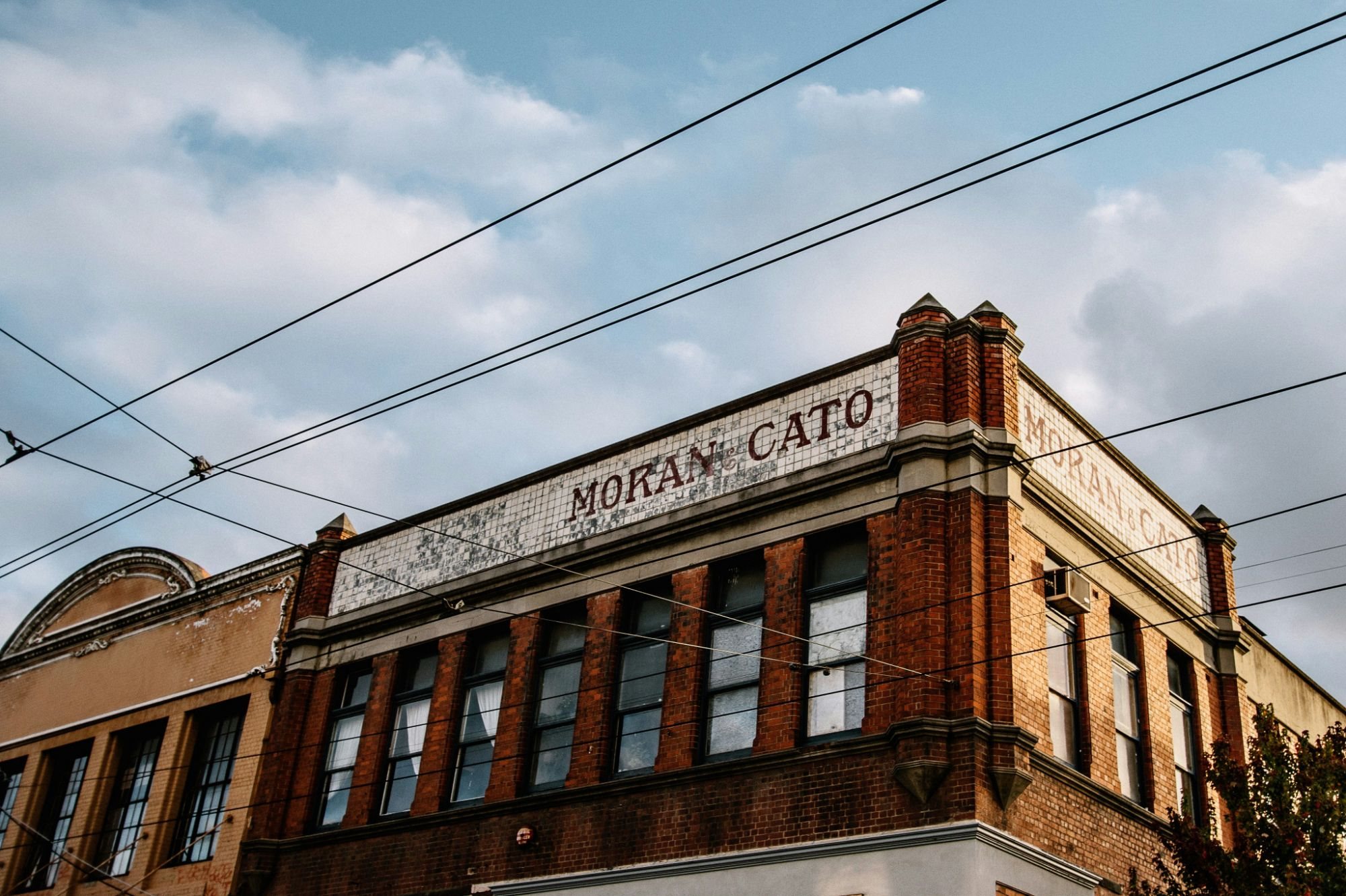 The Moran and Cato building in Fitzroy, Melbourne: red brick and a tiled parapet under tram wires and a cloudy sky
