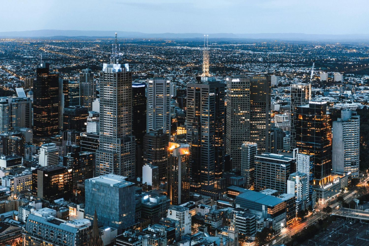 Central Melbourne at dusk from above: office towers with lit windows, the suburbs and ranges beyond